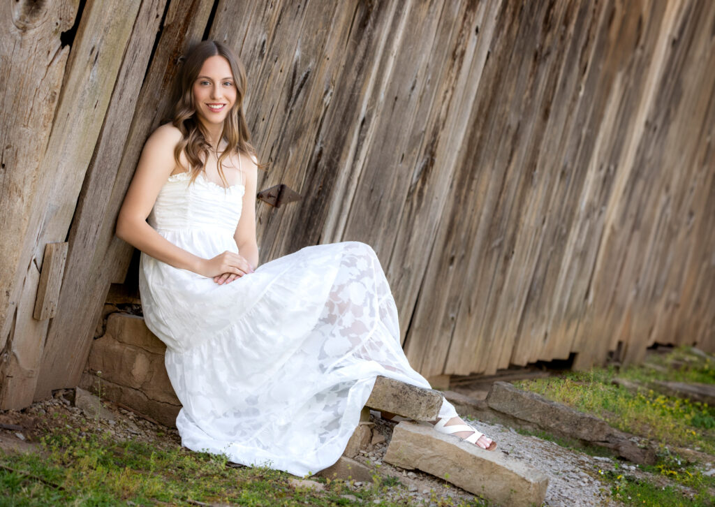 High school senior Spring season brown hair fair complexion white dress sitting against barn in background