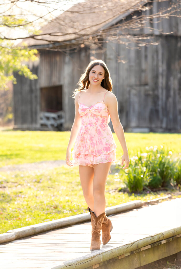 High school senior Spring season brown hair fair complexion cowgirl boots pink floral dress walking across bridge barn in background