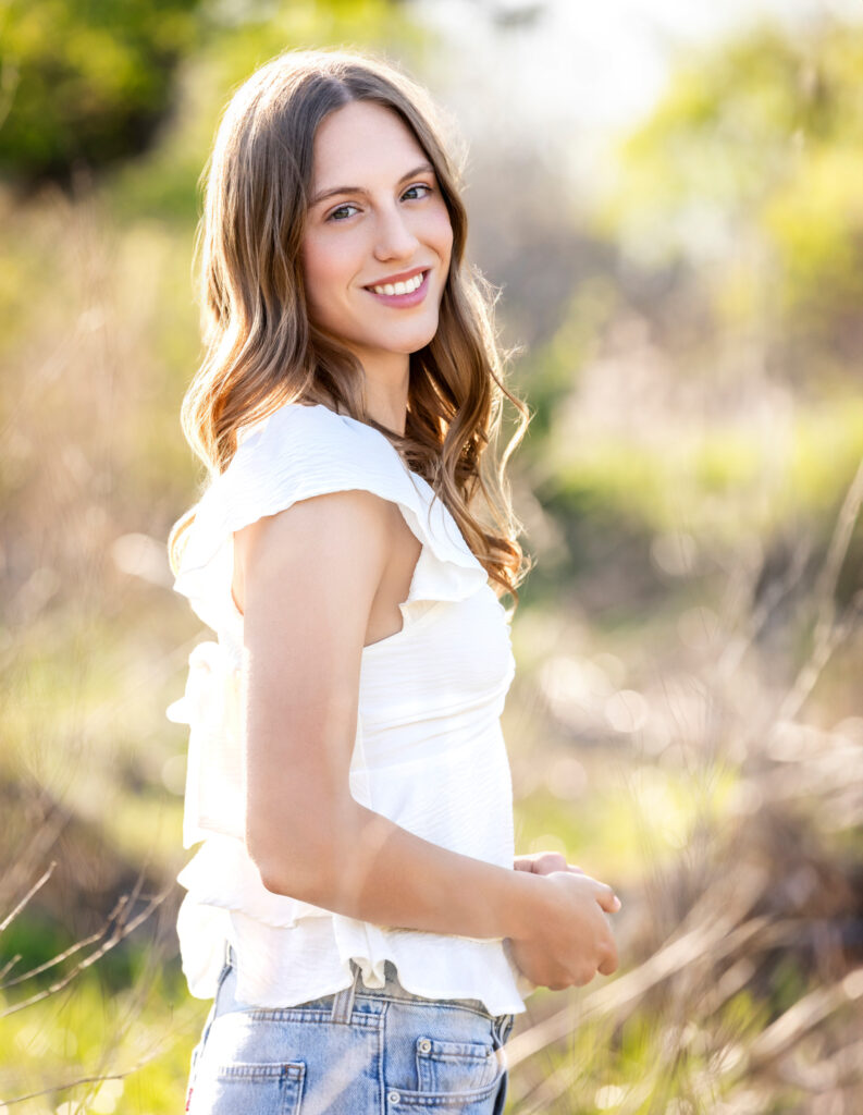 High school senior Spring season brown hair fair complexion in field at sunset hands in front jeans and white shirt