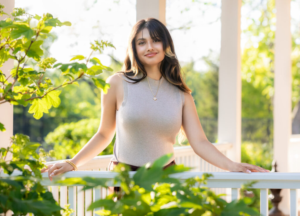 High school senior standing on a front porch in a tan top long dark hair and gold jewelry