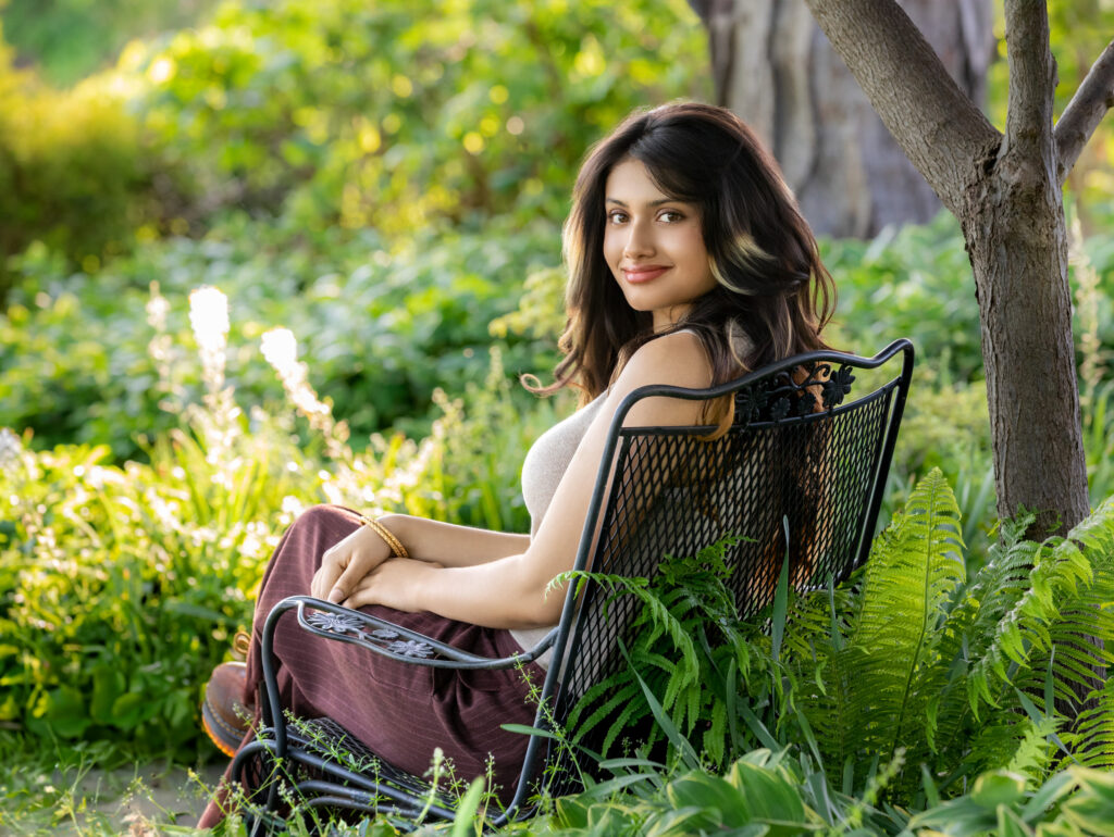 High school senior in a tan top and brown pants with long dark hair sitting on an iron bench with legs and hands crossed looking over shoulder