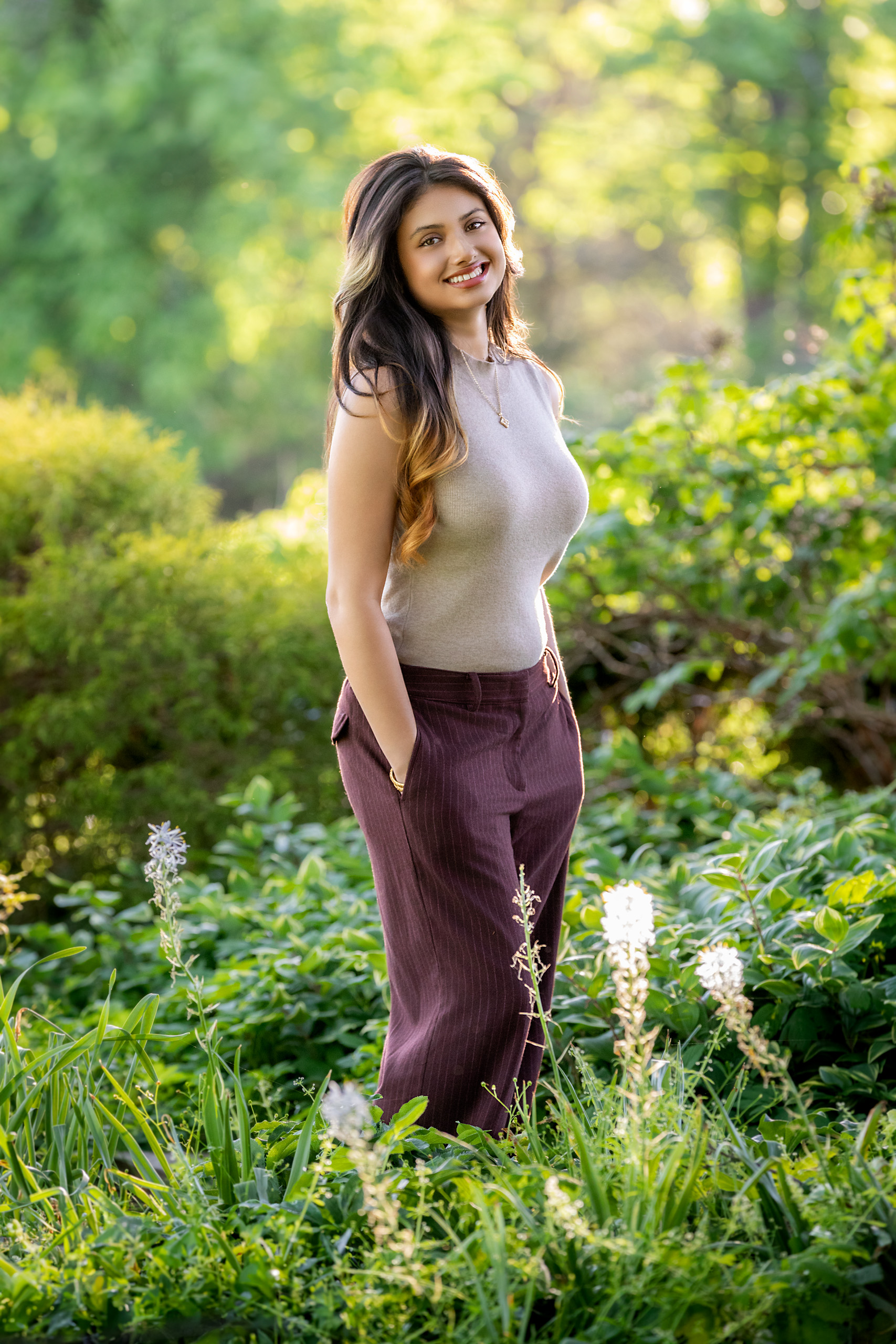 High school senior long dark hair in tan top and brown pants standing in a grassy field