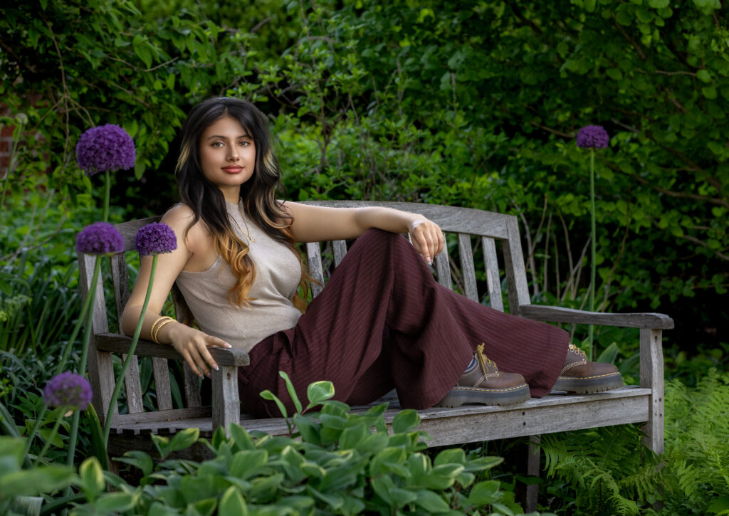 High school senior lounging on a wooden park bench in a tan top and brown pants with long dark hair hand on knee purple flowers
