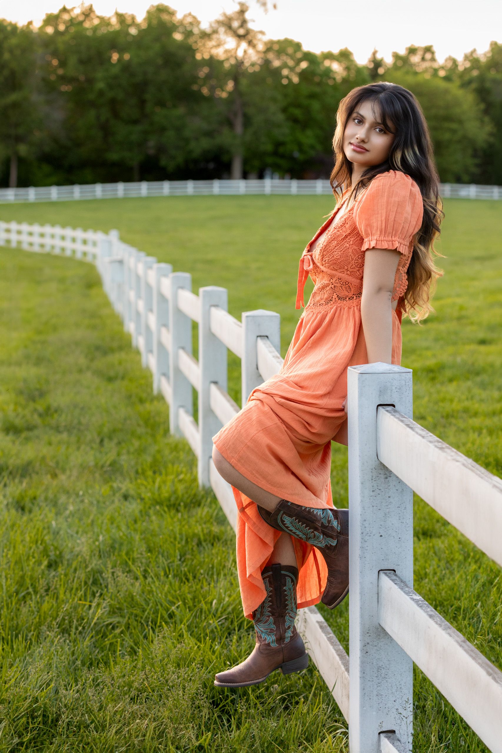 High school senior in an orange dress in cowgirl boots sitting on a fence in a field