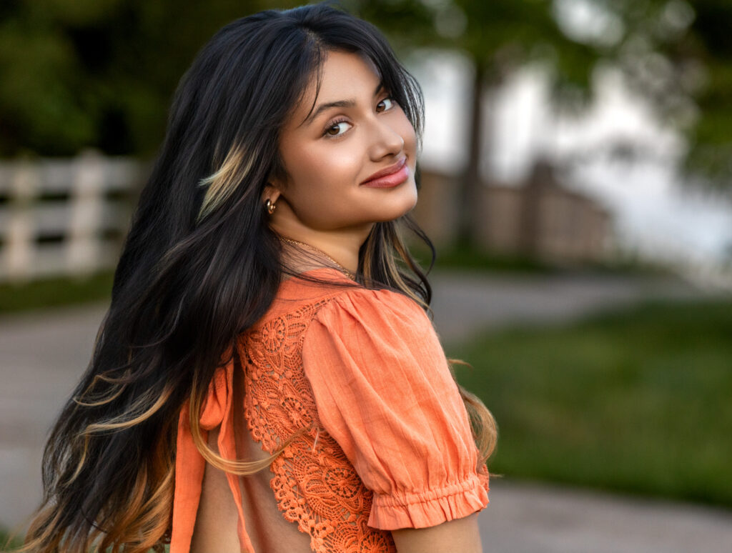 High school senior in an orange dress dark hair with highlights looking over shoulder