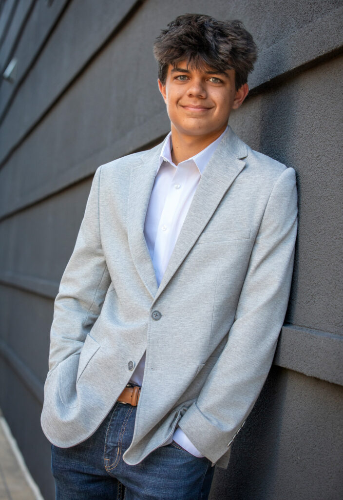 High school senior leaning against a gray building with a light gray blazer and jeans with hands in pockets