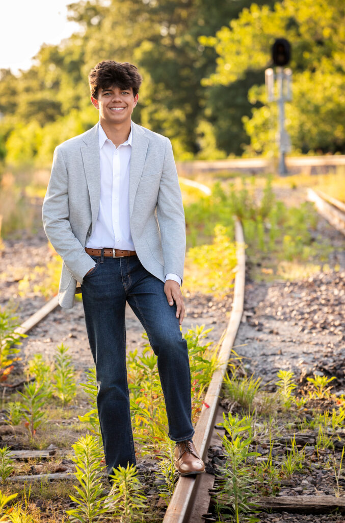 High school senior in gray blazer and jeans standing near railroad tracks one hand in pocket