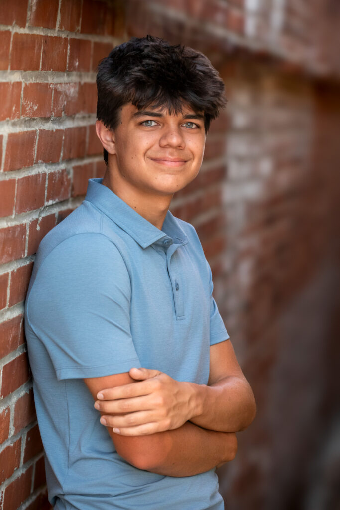 High school senior leaning against a brick wall with a blue polo shirt and light green eyes and dark hair