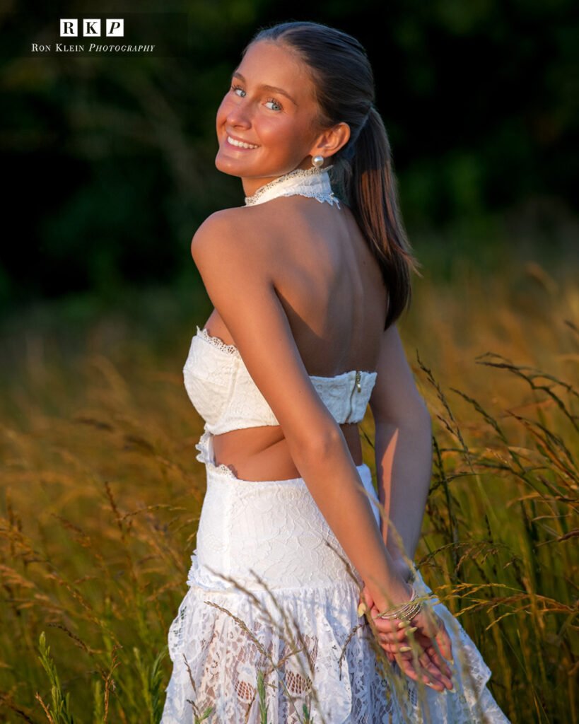 High school senior golden hour white dress in grassy field looking back over shoulder hands behind back