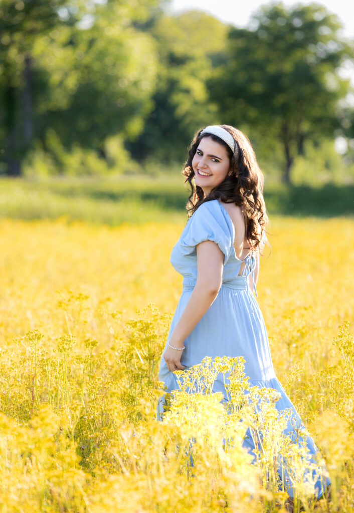 High School Senior Spring Session. Standing in a field of yellow flowers, blue dress, curly hair, looking back.