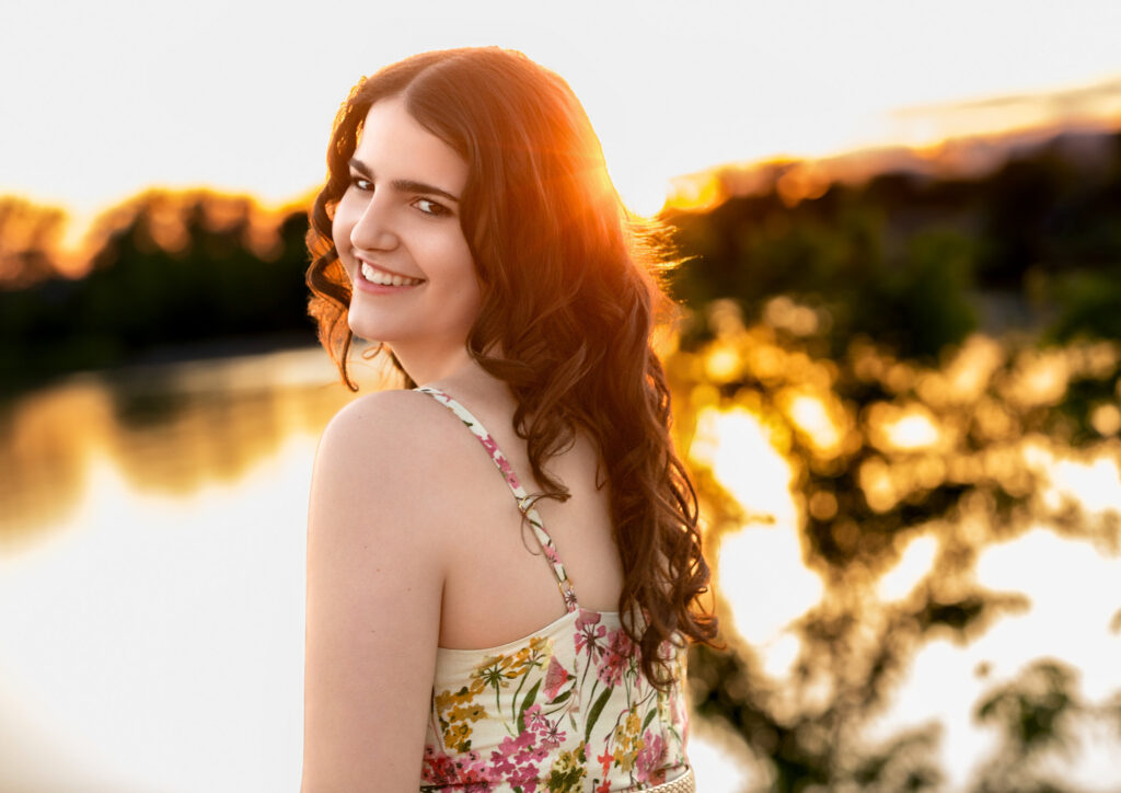 High School Senior Spring Session. Standing by a lake looking over shoulder, floral dress curly hair