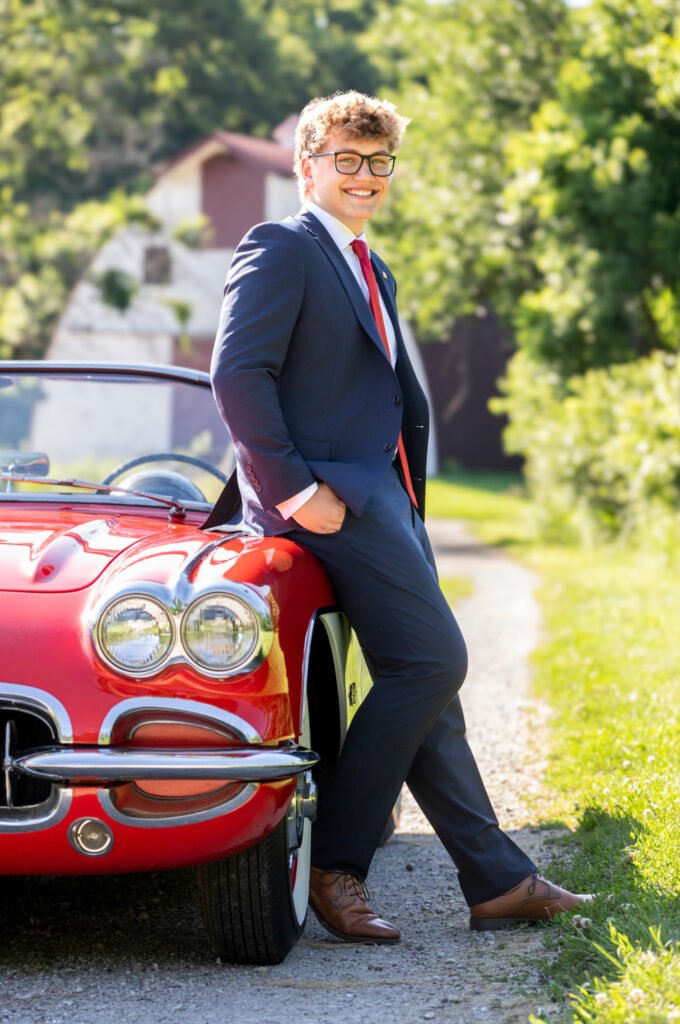 High School Senior red sports car leaning against car wearing a suit and red tie glasses