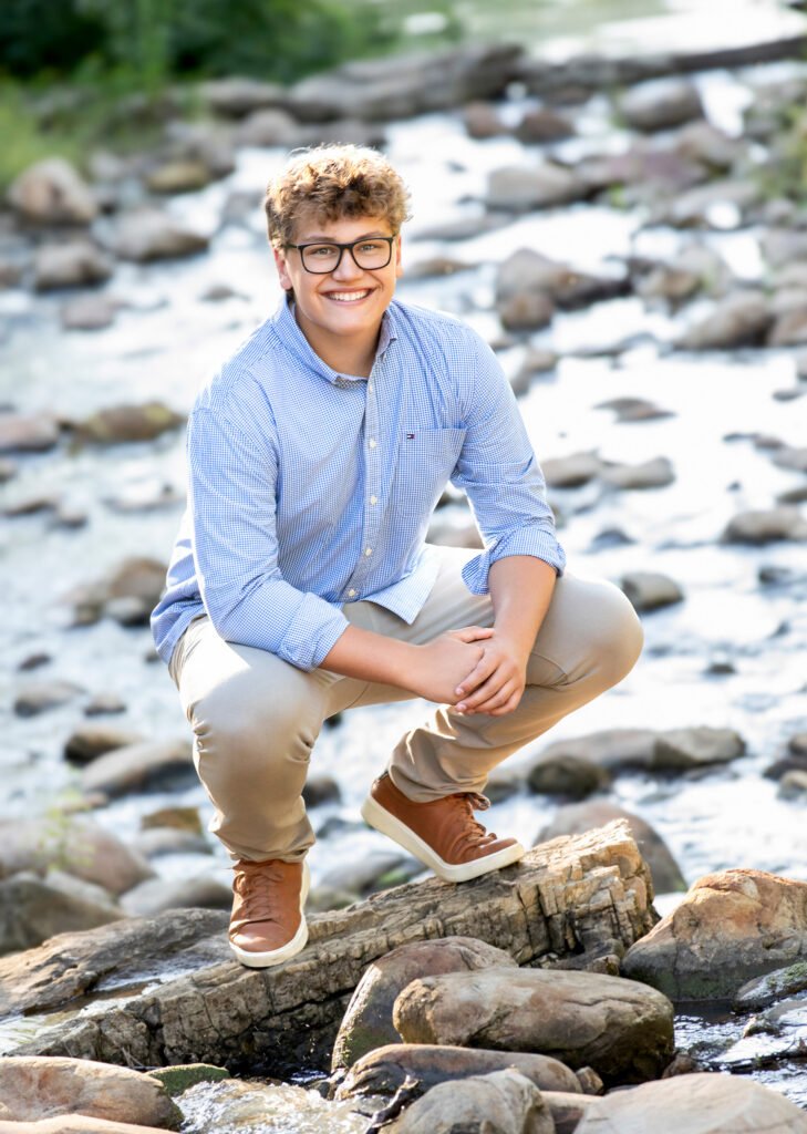High school senior in creek squatting on rocks wearing chinos and an oxford