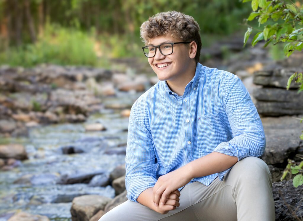 High school senior sitting on a rock in a creek looking in the distance. Wearing glasses and a blue button-up shirt