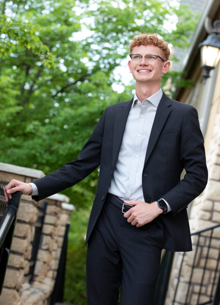 High school senior summer session smiling and looking in the distance wearing a blazer by stone building and metal railings