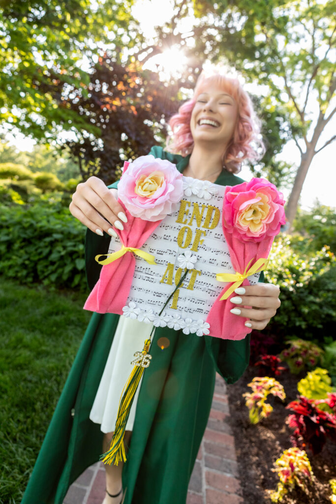 High school senior with pink hair and green robe holding up awards in a park
