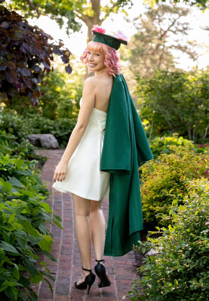 High school senior walking down brick path pink hair green graduation gown draped over shoulder wearing her mortarboard