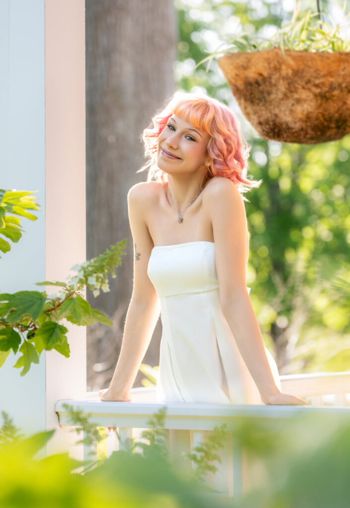 High school senior on porch in white dress and pink hair leaning over railing