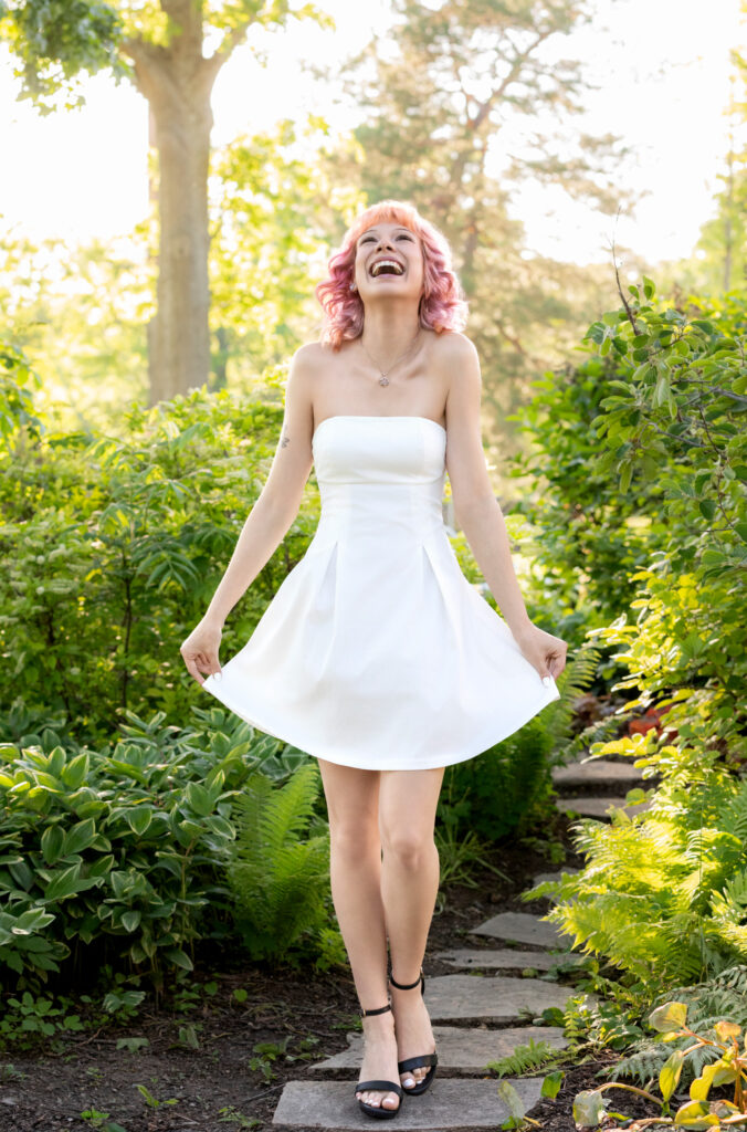 High school senior in white dress and pink hair walking down stone sidewalk looking up laughing hands on dress black sandals