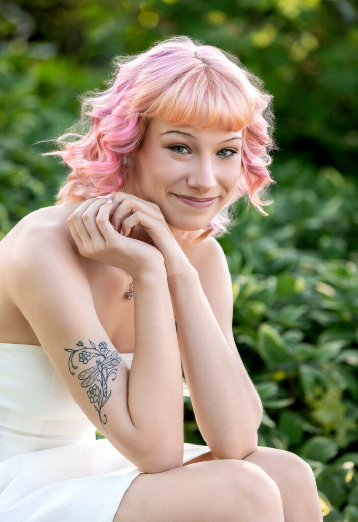 High school senior close up with short pink hair and fair skin arm tattoo sitting in a white dress