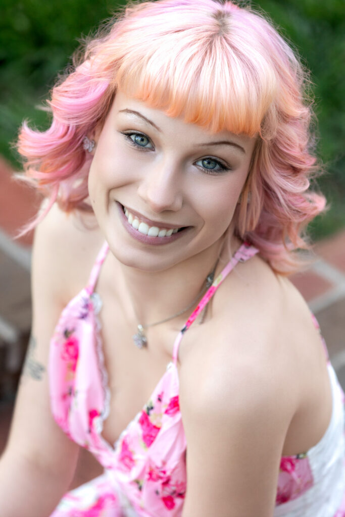 High school senior in a pink floral dress smiling up at the camera pink hair and bangs